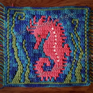 May include: A crocheted square featuring a pink seahorse with white dots, surrounded by green and blue crocheted seaweed. The square has a fringe border and the text "Cynie Birdsong Designs" is visible in the top right and bottom right corners.