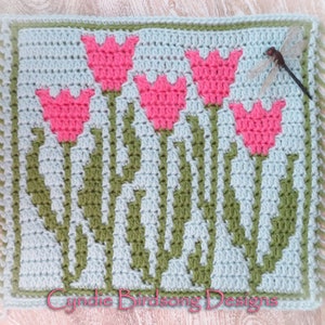 May include: A crocheted square featuring a design of five pink tulips with green stems and leaves on a light blue background. The square has a fringe border and the text "Cynide Birdsong Designs" is at the bottom.