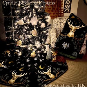 Puede incluir: Una falda de árbol de Navidad de crochet negro y dorado con un diseño de ciervo y una luna creciente. La falda está en el suelo frente a un árbol de Navidad negro con adornos blancos y luces. Una manta de crochet negra y verde con un diseño de ciervo está en una silla en el fondo.