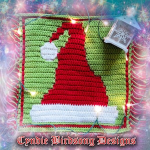 May include: A crocheted square featuring a red and white Santa hat design. The hat is surrounded by a green border and the square is decorated with a string of white lights.