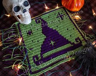 May include: A green and purple crocheted square with a witch's hat design. The square has a fringe border and is surrounded by string lights and a skull.