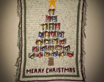 Op de afbeelding: Een gehaakte kerstboom wanddecoratie met een ster bovenop en de woorden "Merry Christmas" onderaan. De boom is versierd met kleine ingepakte cadeautjes.