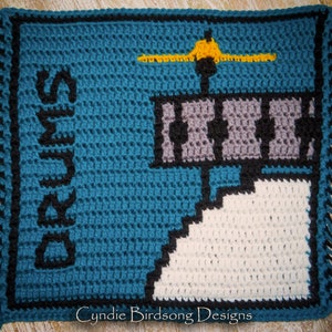 May include: A crocheted square with a blue background, a white and gray building, a yellow airplane, and the text "Sunrise" in black.