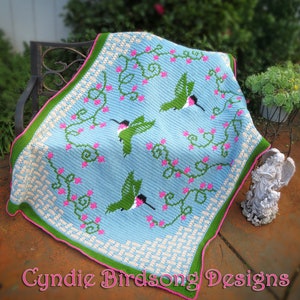 May include: A crocheted throw blanket with a blue background featuring a design of three hummingbirds and green vines with pink flowers. The blanket has a green and white border.