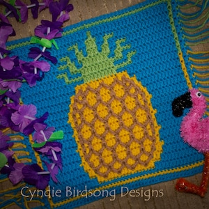 May include: A crocheted pineapple and flamingo design with a blue background and yellow fringe. The design is on a crocheted blanket with a purple flower garland in the corner. The text "Cynthie Birdsong Designs" is at the bottom of the image.