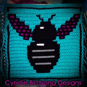 Puede incluir: Un cuadrado tejido a crochet con una abeja negra y blanca con alas moradas. La abeja está sobre un fondo turquesa. El cuadrado tiene un borde negro y un borde de flecos. El texto "Cynide Birdsong Designs" está en la parte inferior del cuadrado.