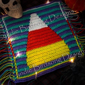 May include: A handmade crochet pillow featuring a candy corn design. The pillow has a white, red, and yellow candy corn in the center, with a striped border in teal, purple, and green. The edges have colorful fringe. Text on the pillow reads "Cyndie Birdsong Designs."