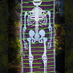 May include: A crocheted skeleton design in white, set against a backdrop of purple, green, and black horizontal stripes. The skeleton's skull, rib cage, and limbs are clearly visible. The text "Cyndie Birdsong Designs" is at the bottom.