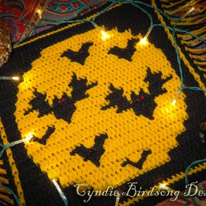 Può includere: Un quadrato all'uncinetto fatto a mano a tema Halloween, con una luna gialla e pipistrelli neri con occhi rossi. Il quadrato ha un bordo nero. Il testo "Cyndie Birdsong Designs" è visibile. Sono presenti anche luci decorative e una statuina di gatto nero.
