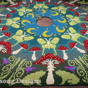 Puede incluir: Una falda de árbol de crochet verde, azul y amarilla con un diseño de mandala que presenta hongos, mariposas y lunas crecientes. "Cynie Birdsong Designs" está escrito en la esquina inferior izquierda.