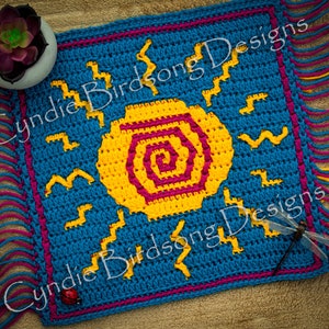 May include: A square, crocheted sun design in shades of blue, yellow, and magenta. The sun has a spiral center and is surrounded by zigzag and bird-like shapes. The edges are trimmed with fringe and the text "Cyndie Birdsong Designs" is visible.