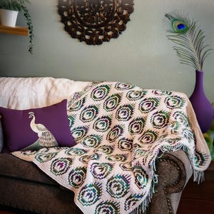 May include: A cozy living room scene features a brown sofa adorned with a cream-colored throw and a decorative pillow with a peacock design. A cream-colored blanket with a peacock feather pattern is draped over the sofa. The text "ENCHANTED PEACOCK PLUMES" is visible.