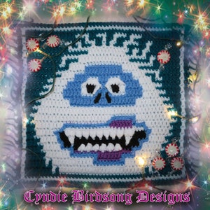 May include: A crocheted square featuring a snowman with a blue face, white body, and purple mouth. The snowman is surrounded by a green border with white candy canes. The text "Cynie Birdsong Designs" is at the bottom of the square.