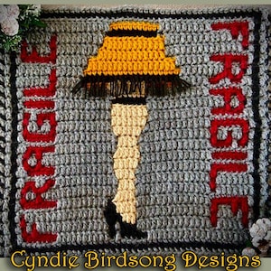 May include: A crocheted rug with a gray background and red fringe. The center of the rug features a crocheted image of a lampshade with legs, and the words "LEG LAMP" and "FRUGAL" are crocheted on either side of the image.