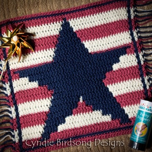 May include: A crocheted patriotic-themed square featuring a navy blue star on a background of red and white stripes. The square has a fringed border and the text "Cyndie Birdsong Designs". A gold pinwheel and a party popper are also in the image.