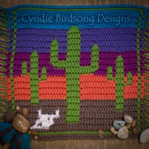 May include: A crocheted wall hanging depicting a desert scene with three green cacti and a white skull. The background is a multi-coloured striped pattern of blue, purple, orange, and brown. The text "Cynjie Birdsong Designs" is at the top of the hanging.
