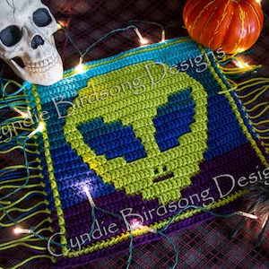 May include: A green, blue, and purple crocheted skull with fringe on all sides. The skull is surrounded by string lights and a small pumpkin. The text "Cynthie Birdsong Designs" is visible on the crocheted skull.