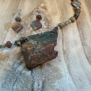 May include: A necklace and earrings set featuring a large, irregularly shaped pendant made of brown, green, and red jasper. The pendant is attached to a silver chain with smaller jasper beads. The earrings are silver with jasper drops.