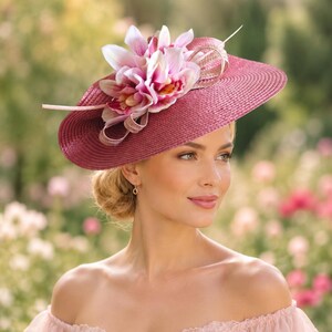 May include: A wide-brimmed magenta straw hat adorned with a cluster of light pink and white artificial lily flowers and decorative ribbon loops. The hat features a textured, woven straw construction and is positioned atop the head.