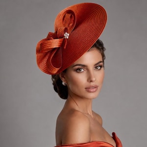 May include: A burnt orange fascinator hat featuring a wide, circular brim and a large, structured bow detail on the side. The center of the bow is accented with a small cluster of white faux pearls. The hat is crafted from a textured, woven material.