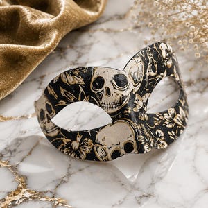 May include: A black masquerade mask featuring a repeating pattern of off-white skulls and gold-toned floral accents. The mask has two eye openings and a curved silhouette, showcasing a detailed graphic print across the entire front surface.