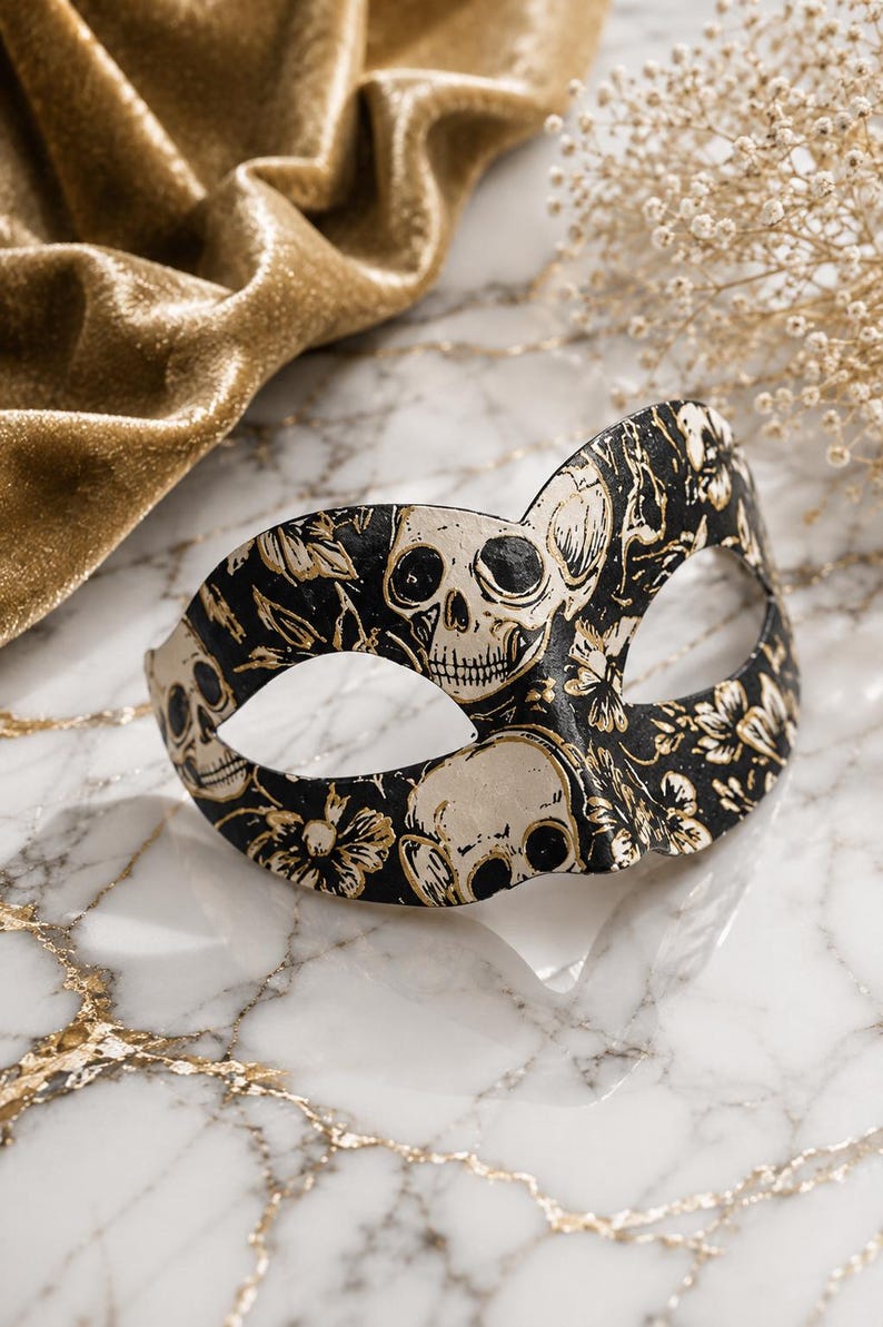 May include: A black masquerade mask featuring a repeating pattern of off-white skulls and gold-toned floral accents. The mask has two eye openings and a curved silhouette, showcasing a detailed graphic print across the entire front surface.
