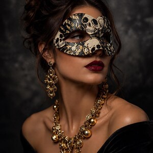 May include: A black and cream masquerade mask featuring a detailed skull and floral pattern, paired with ornate gold-toned drop earrings and a matching gold-toned statement necklace with spherical accents and intricate filigree detailing.