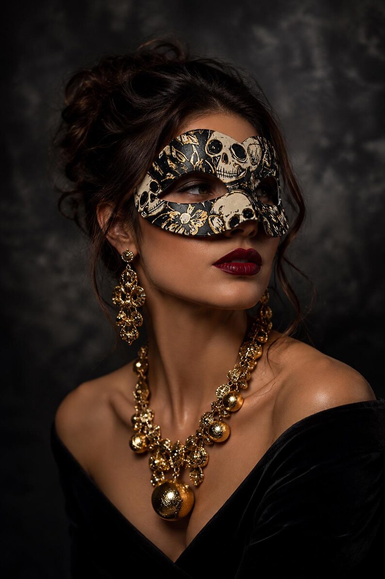 May include: A black and cream masquerade mask featuring a detailed skull and floral pattern, paired with ornate gold-toned drop earrings and a matching gold-toned statement necklace with spherical accents and intricate filigree detailing.