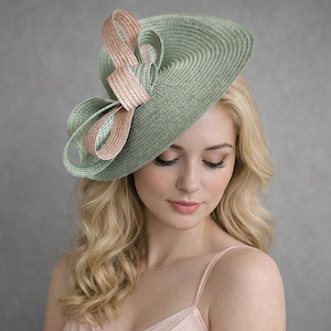 May include: A pale green hat with a pink bow. The hat is made of woven material and has a wide brim. The bow is a lighter pink colour. The hat is worn by a person with blonde hair.