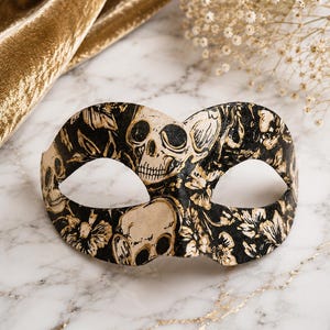 May include: A masquerade mask featuring a black background with a repeating pattern of cream-colored skulls and floral motifs. The mask has two eye openings and a curved shape designed to cover the upper portion of the face with detailed art.