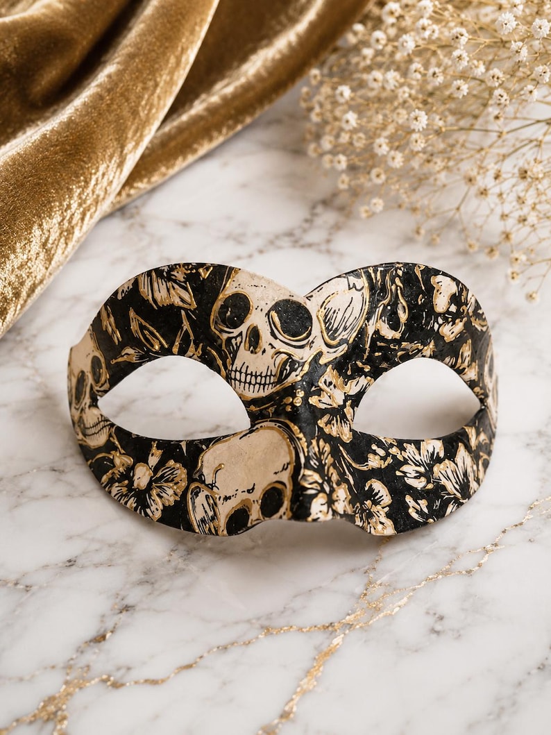 May include: A masquerade mask featuring a black background with a repeating pattern of cream-colored skulls and floral motifs. The mask has two eye openings and a curved shape designed to cover the upper portion of the face with detailed art.