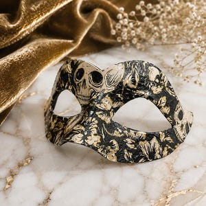 May include: A masquerade mask featuring a black base with a gold floral pattern and skull motifs. The mask has two eye openings and a contoured shape designed to cover the upper portion of the face, showcasing a detailed, textured surface finish.
