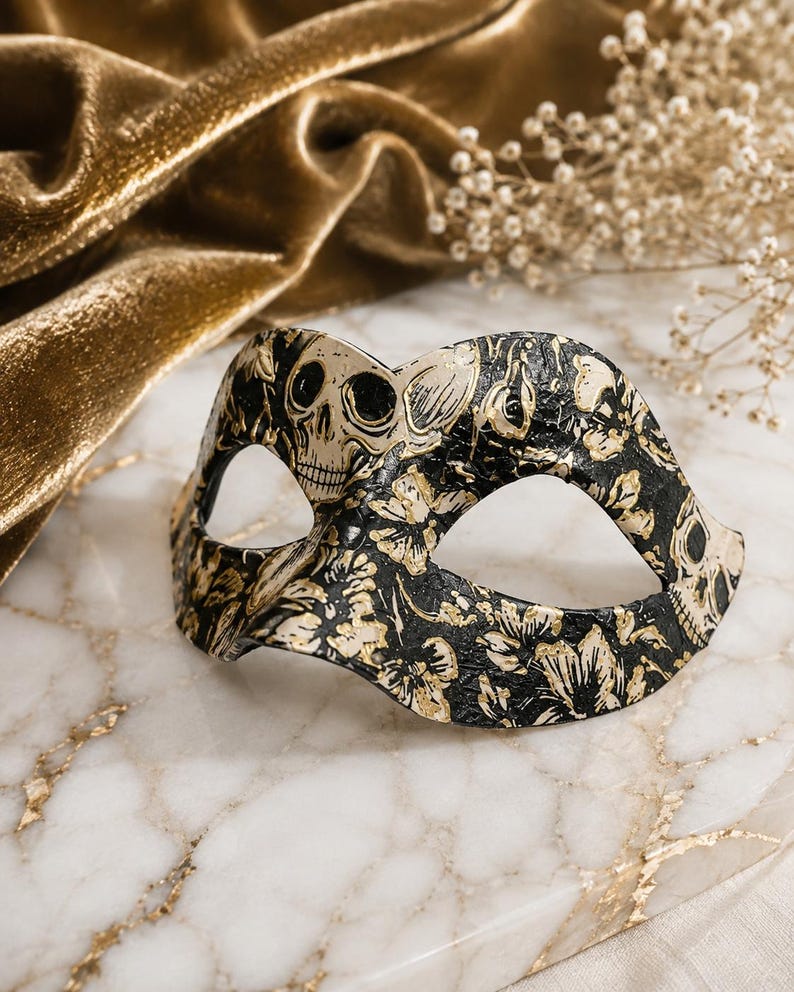 May include: A masquerade mask featuring a black base with a gold floral pattern and skull motifs. The mask has two eye openings and a contoured shape designed to cover the upper portion of the face, showcasing a detailed, textured surface finish.