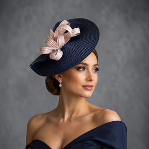 Op de afbeelding: Een marineblauwe strooien fascinatorhoed met een brede, ronde rand en een decoratieve, meervoudig geluste strik in een zachte, lichtroze tint. De hoed is schuin geplaatst, wat de textuur van het vlechtwerk en de gestructureerde strik benadrukt.