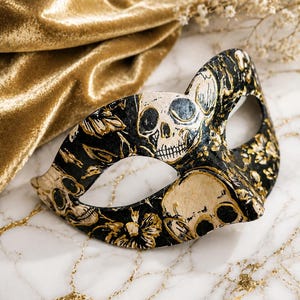 May include: A masquerade mask featuring a black base with a detailed gold and cream-colored pattern of skulls and floral motifs. The mask has two eye openings and a contoured shape designed to cover the upper portion of the face.
