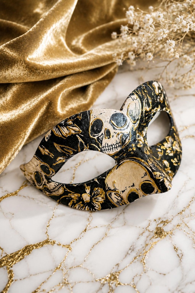 May include: A masquerade mask featuring a black base with a detailed gold and cream-colored pattern of skulls and floral motifs. The mask has two eye openings and a contoured shape designed to cover the upper portion of the face.