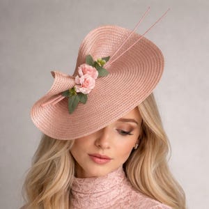 Könnte beinhalten: Ein hellrosa Hut mit breiter Krempe und floraler Verzierung. Der Hut hat eine geschwungene Krempe und ist mit rosa Rosen und grünen Blättern verziert. Zwei dünne, rosa federartige Akzente ragen aus dem Hut. Ein modisches Accessoire.