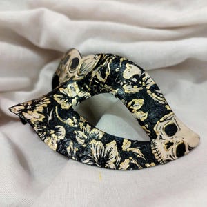 May include: A black masquerade mask featuring a detailed pattern of white and gold floral motifs interspersed with small skull illustrations. The mask has a pointed, almond-shaped design with a central eye opening and a textured surface finish.