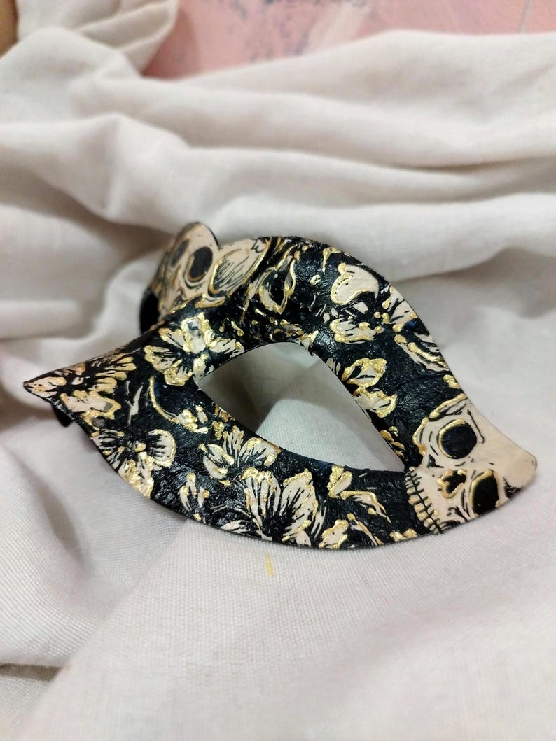 May include: A black masquerade mask featuring a detailed pattern of white and gold floral motifs interspersed with small skull illustrations. The mask has a pointed, almond-shaped design with a central eye opening and a textured surface finish.