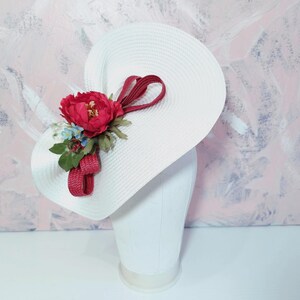 May include: A white wide-brimmed hat featuring a decorative arrangement on the side. The embellishment consists of a large dark pink peony, small light blue flowers, green leaves, and a looped dark pink textured ribbon accent.