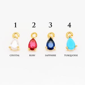 Peut inclure: Quatre breloques en forme de goutte d'or avec des pierres précieuses de différentes couleurs. Les breloques sont numérotées de 1 à 4. La breloque 1 est en cristal, la breloque 2 est en rubis, la breloque 3 est en saphir et la breloque 4 est en turquoise.