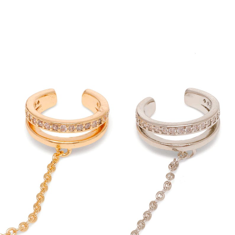May include: Two ear cuffs, one gold-toned and one silver-toned, each featuring a double-band design with a row of small clear stones embedded in the upper band and a delicate metal chain hanging from the lower portion of the cuff structure.