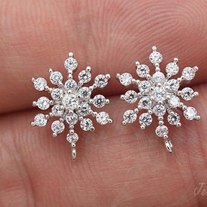 May include: A pair of silver-toned snowflake stud earrings, each adorned with multiple clear, round-cut gemstones. The earrings have a post and loop for attachment. The design is intricate and detailed, resembling a winter theme.