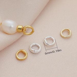 以下が含まれることがあります： 金と銀色の丸カンと真珠を含む、ジュエリーパーツのクローズアップ。丸カンの直径は約5mmです。背景は薄いピンク色の生地です。