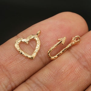 Può includere: Due ciondoli in metallo dorato, uno a forma di cuore e l'altro a forma di freccia. Entrambi i ciondoli hanno una superficie testurizzata.