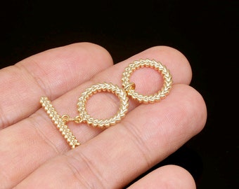 May include: A gold-coloured toggle clasp with a textured design. The clasp features two circular rings and a bar, all with a repeating pattern. The jewellery finding is shown against a black background.