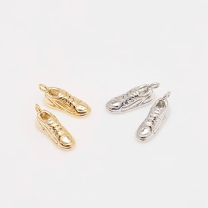 May include: Four small sneaker charms, two in gold and two in silver, are displayed against a white background. The charms are detailed with laces and soles, perfect for jewelry making or crafting projects.
