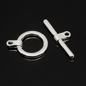 Puede incluir: Cierre de palanca plateado con un anillo redondo y una barra recta. El anillo y la barra tienen una ranura decorativa.