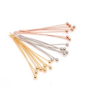 Puede incluir: Una colección de alfileres de cabeza en colores oro, plata y cobre. Cada alfiler tiene una pequeña bola en el extremo. Estos alfileres se utilizan para la fabricación de joyas y manualidades.