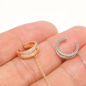 May include: Two ear cuffs with attached chains, one in a rose gold tone and one in a silver tone. Each cuff features a double-band design with a row of small, clear, sparkling stones embedded along the outer band, connected to a thin metal chain.